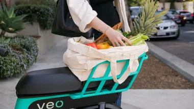 Close-up on back basket of teal and black Veo bike share bike with woman's hands reaching into canvas bag full of produce.